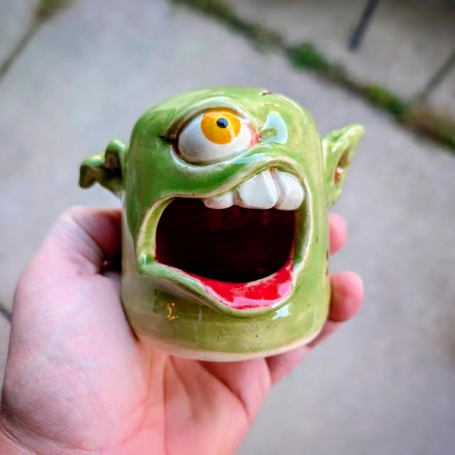 A green one-eyed goblin mug with a toothy grin
